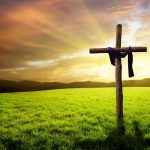 A wooden cross draped in a purple robe, standing in a grassy field beneath a beautiful sunrise.